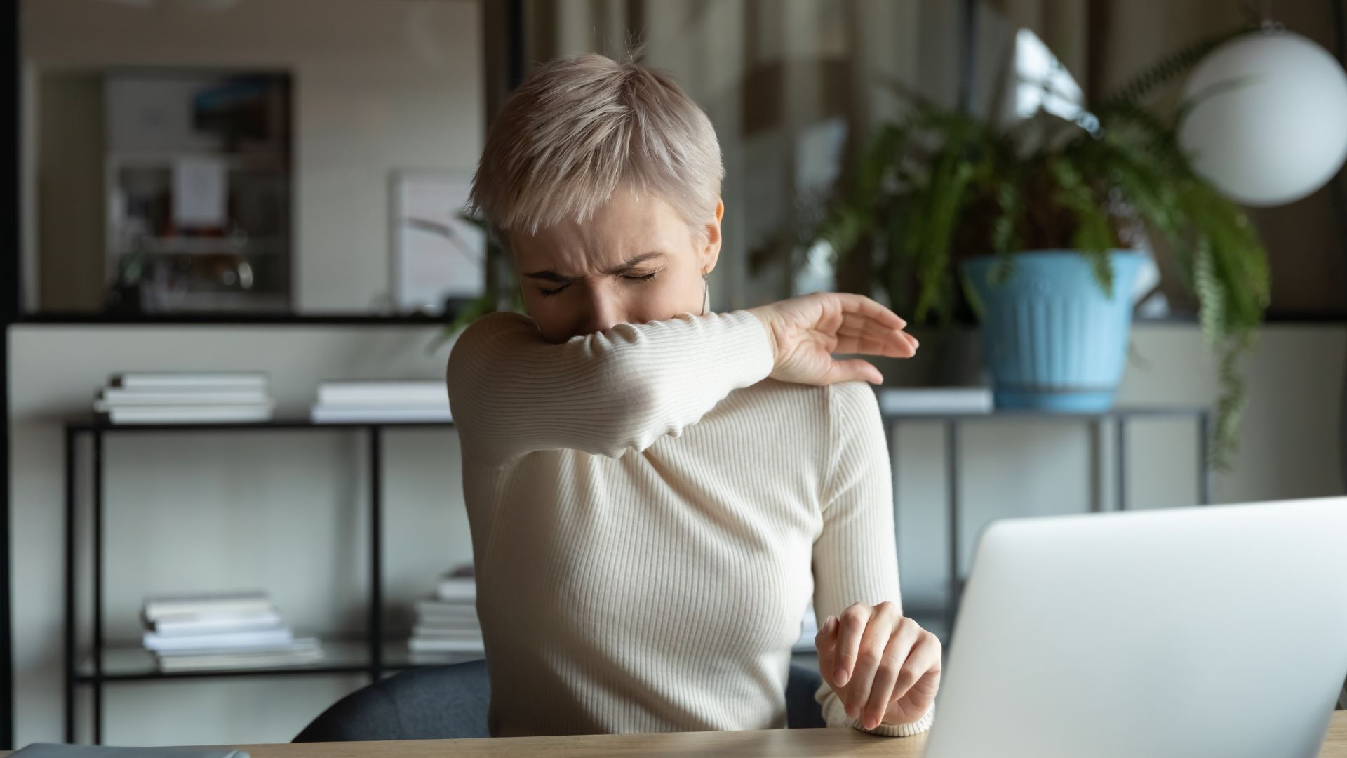 Woman at desk, sneezing into her arm