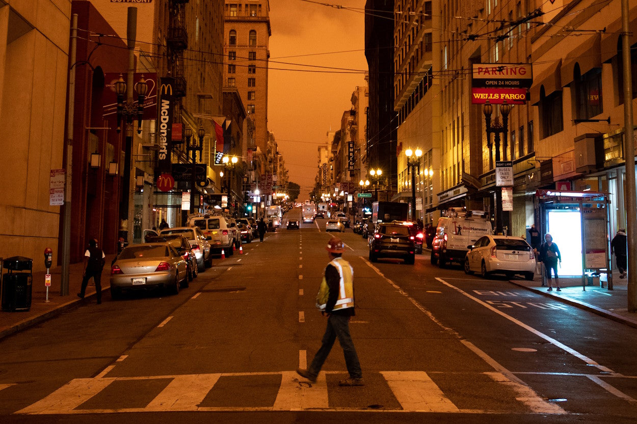 Man walking across street with sky colored orange by wildfire smoke